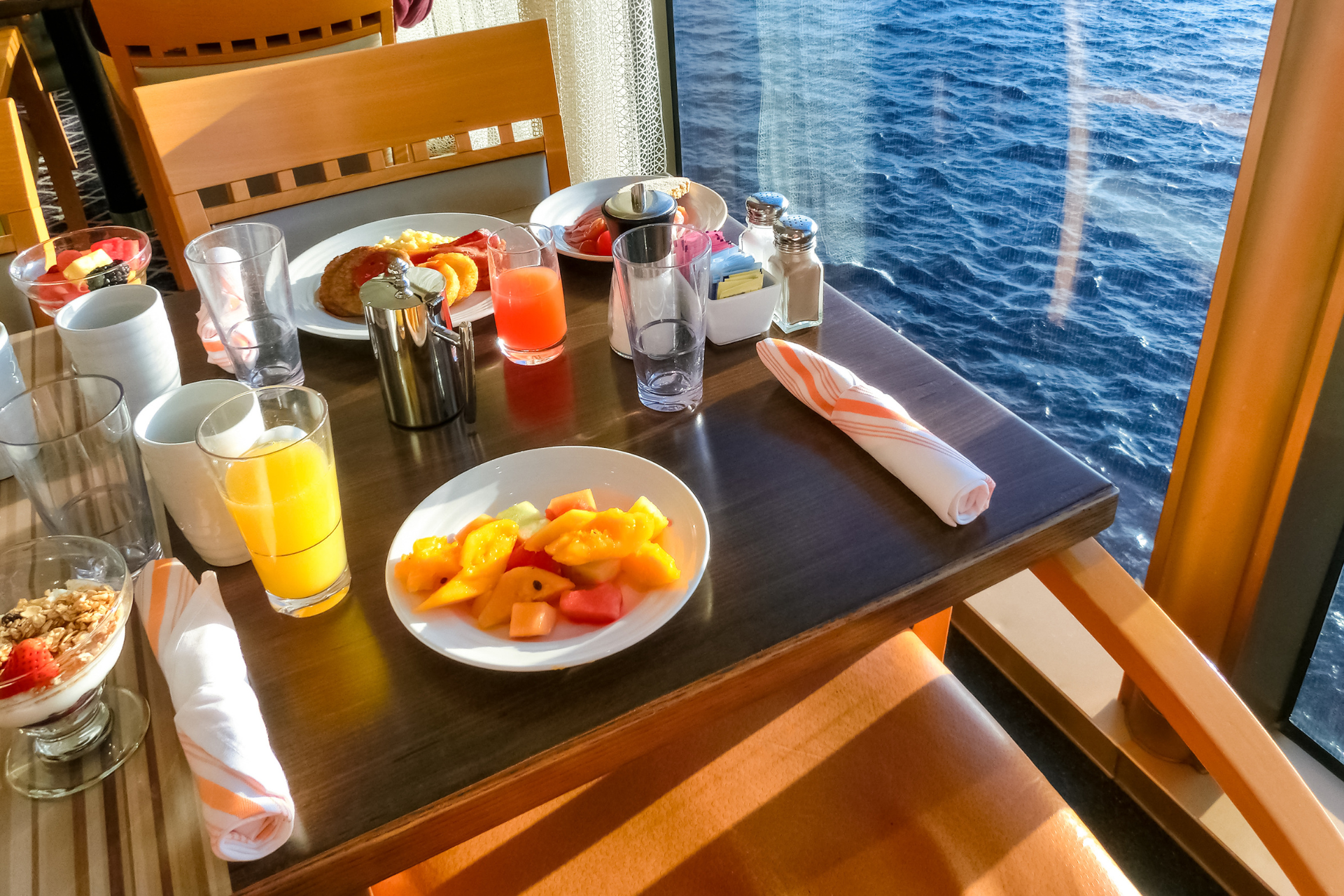 A breakfast table set with fresh fruits, juices, and pastries, overlooking the ocean through a large window, capturing the essence of culinary cruising adventures.