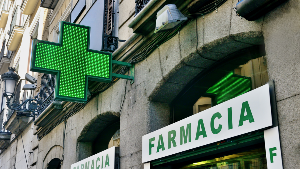 An illuminated green cross sign outisde a building make Farmacia.  This is a typical sign for pharmacies in Europe.  Getting medical help and drug refills can be expensive without travel insurance.