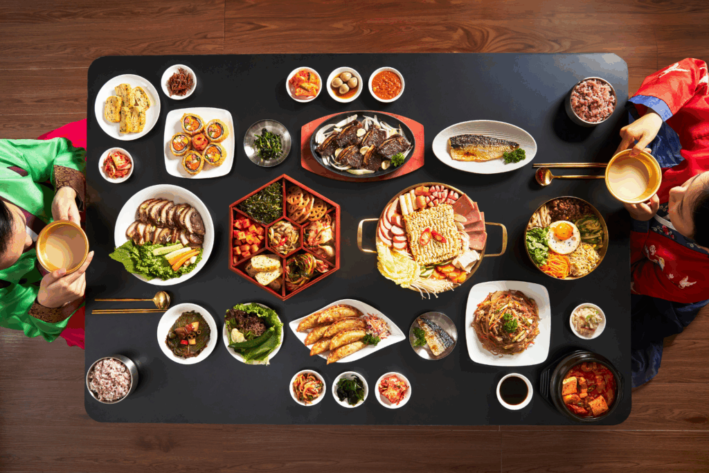 Displayed is  an overhead shot of beautifully displayed traditional Korean culinary dishes on a black table with a person on each end holding a bowl of broth.